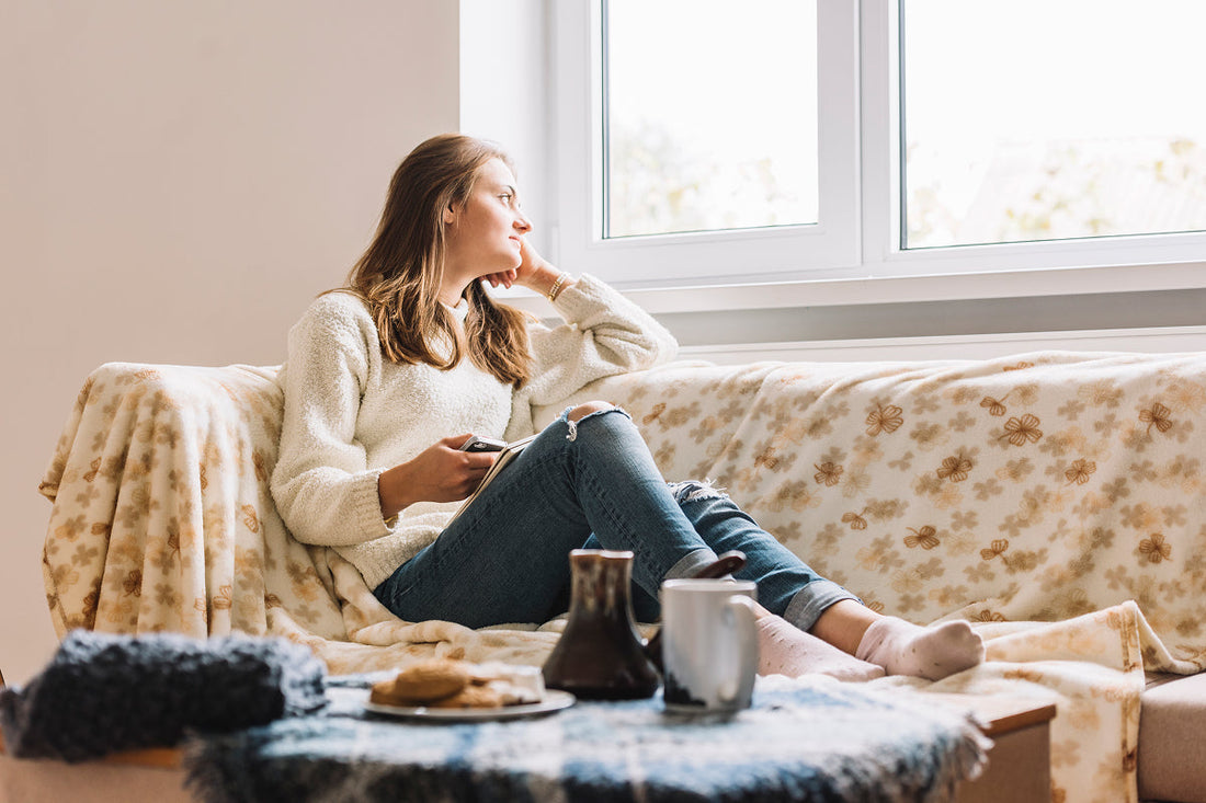 young woman relaxing with book on sofa looking outside window with tea and biscuits on a table