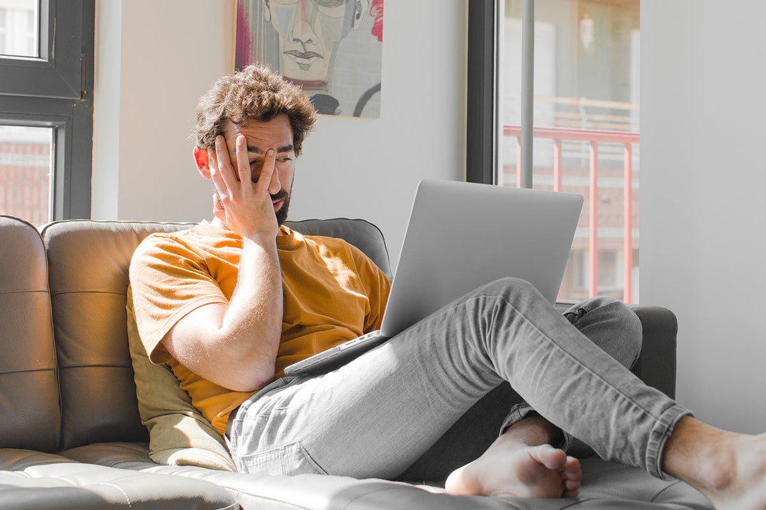 Man sitting on couch with laptop on lap holding hand over head